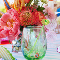 A Kim Seybert Gala Stemless Tumbler from the Set of 4, filled with a green drink and decorative straw, rests on a striped tablecloth surrounded by vibrant pink, orange, and red flowers in glass vases.