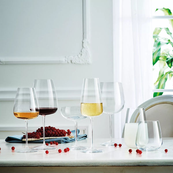 Elegant Luigi Bormioli Talismano Bar Collection Sets wine glasses, filled with red, white, and rosé wine, are artfully arranged on a white table with scattered red berries, a blue napkin, and two glasses of milk in a bright classic interior.