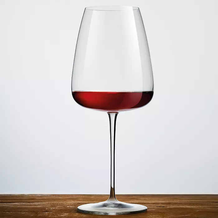 A Luigi Bormioli Talismano Bar Collection Set wine glass with a thin stem rests on a wooden surface, partially filled with red wine, against a plain light grey background, showcasing elegant Italian craftsmanship.