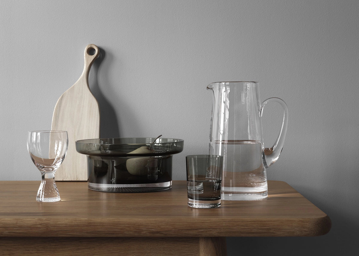 A wooden table with a Kosta Boda Limelight Jug, a crystal glass, a water glass, a goblet, a dark glass bowl of fruit, and a light wooden cutting board set against a gray wall.