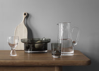 A wooden table with a Kosta Boda Limelight Jug, a crystal glass, a water glass, a goblet, a dark glass bowl of fruit, and a light wooden cutting board set against a gray wall.