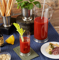 A glass and a pitcher of Bloody Mary with celery, served with breadsticks, cheese, olives, salami, and a lemon wedge on a blue tablecloth—presented in the Italian artisan-crafted MATCH Martini Crystal Pitcher by MATCH.