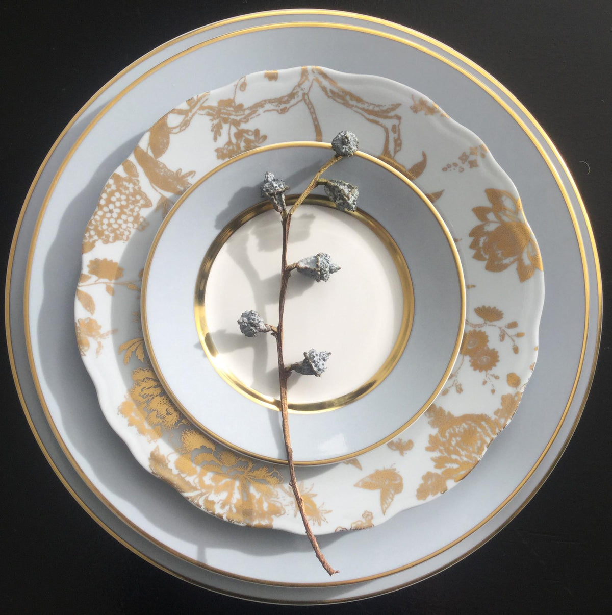 A stem with frosted berry-like decorations rests on a stack of three Royal Limoges Recamier Grey & Gold dinner plates, featuring a pale palette and intricate gold detailing.