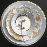 A stem with frosted berry-like decorations rests on a stack of three Royal Limoges Recamier Grey & Gold dinner plates, featuring a pale palette and intricate gold detailing.