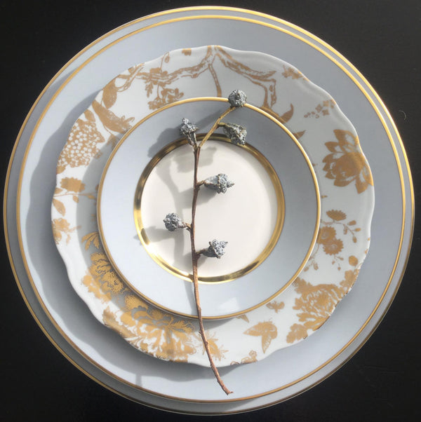 A stem with frosted berry-like decorations rests on a stack of three Royal Limoges Recamier Grey & Gold dinner plates, featuring a pale palette and intricate gold detailing.