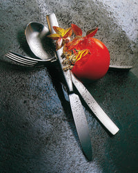 A tomato and leafy herbs rest on Mepra’s Mediterranea Flatware 5 Piece Setting—arranged in a cross on a wet, dark textured surface with scattered water droplets and pepper.