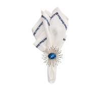 The Kim Seybert Filament Napkin White & Navy Set of 4 features white linen napkins with navy stitched edges, elegantly folded and secured by a round silver sunburst napkin ring with a glossy blue center.