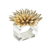 The Stellar Napkin Ring Gold Set of 4 by Kim Seybert features a clear rectangular Lucite design topped with a large gold flower, its spiky petals arranged in a striking circular pattern.