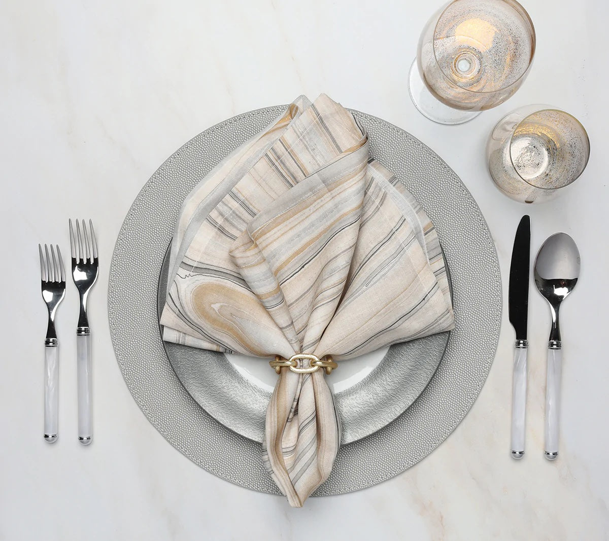 Elegant table setting features a gray and white plate, striped napkin with a gold ring, cutlery, and glasses—all arranged on the Kim Seybert Shagreen Placemat Elephant Grey Set of 4 atop a white surface.