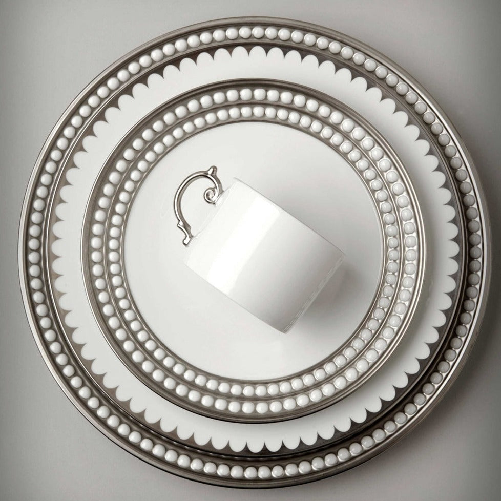 A white teacup rests atop L'Objet Perlée Platinum Dinnerware, each piece featuring platinum rims and silver beaded details, viewed from above on a light gray background.