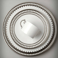 A white teacup rests atop L'Objet Perlée Platinum Dinnerware, each piece featuring platinum rims and silver beaded details, viewed from above on a light gray background.