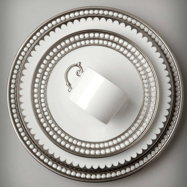 A white teacup rests atop L'Objet Perlée Platinum Dinnerware, each piece featuring platinum rims and silver beaded details, viewed from above on a light gray background.
