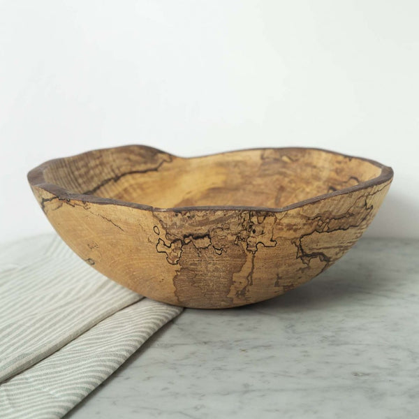 The Peterman Spalted Maple Oval Wood Bowl 18", featuring unique dark grain patterns, rests on a marble surface beside a folded light-colored cloth.