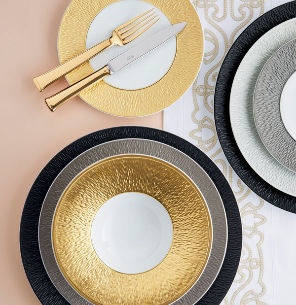 Elegant table setting featuring Raynaud Mineral Irise Dinnerware in Black with textured rims, gold cutlery on a gold-rimmed plate, and a white and silver decorative table runner underneath.
