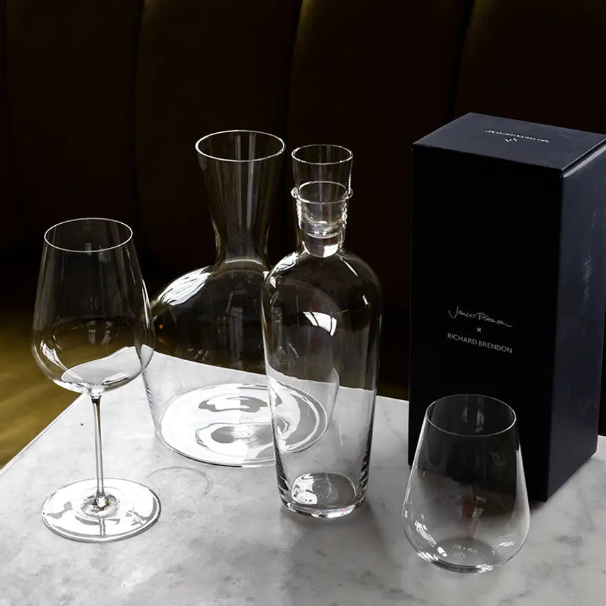 The Richard Brendon Jancis Robinson Young Wine Decanter, a stemless glass, carafe with stopper, wine glass, and a black rectangular box are displayed on a white marble surface.
