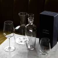 The Richard Brendon Jancis Robinson Young Wine Decanter, a stemless glass, carafe with stopper, wine glass, and a black rectangular box are displayed on a white marble surface.