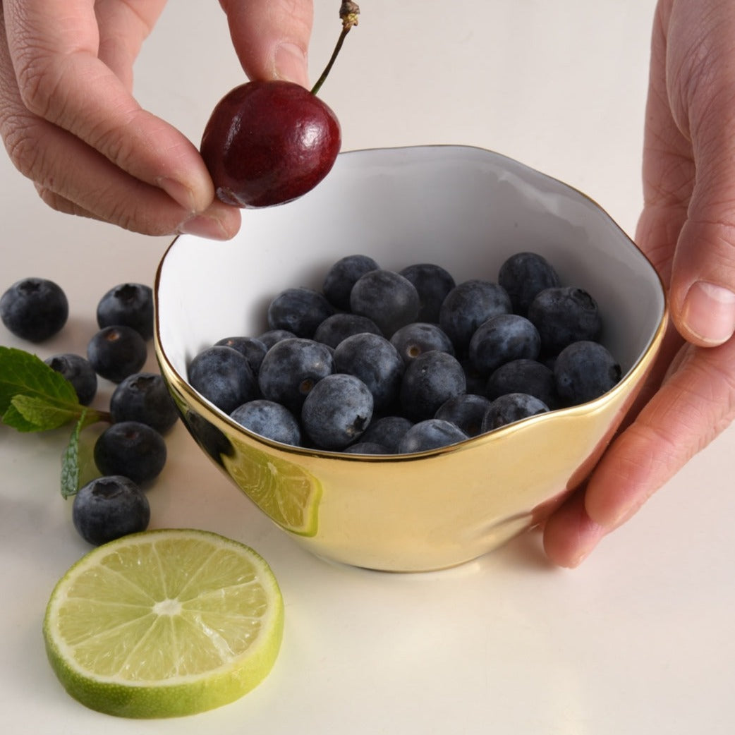 A person holds a PAMPA BAY Moonlight Snack Bowl filled with blueberries in one hand and a cherry above the bowl in the other, with a slice of lime and a mint sprig on the white surface beside it.