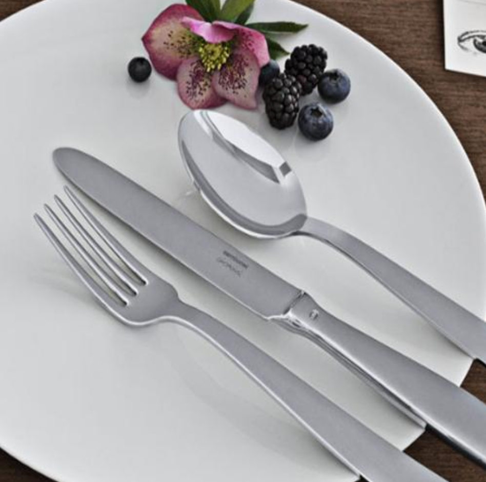 A white plate with the Sambonet Gio Ponti Flatware 5 Piece Setting—fork, knife, and spoon—neatly arranged beside a pink flower, blackberries, blueberries, and greenery on a brown table with a business card in view.