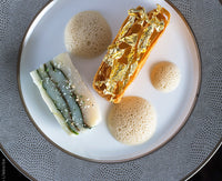 A gourmet dish is served on Bernardaud’s Sauvage Grey & Platinum Limoges porcelain, featuring a gold-topped rectangular pastry, layered vegetable terrine, and three foam pools, all elegantly arranged on a glass charger.
