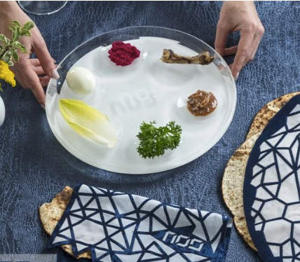 A person arranges symbolic foods—egg, parsley, horseradish, charoset, bone, and lettuce—on the Apeloig Acrylic Seder Plate, placed on a blue tablecloth beside matzah and a napkin with a geometric pattern.