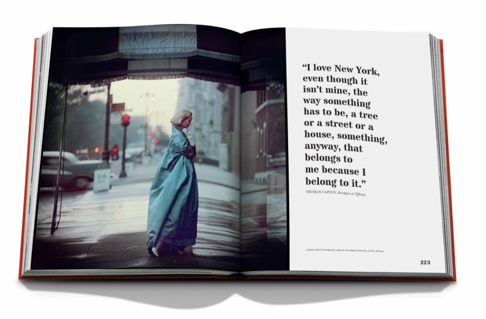 The Assouline book "New York By New York" is open to a spread with a striking image of someone in a long blue coat on a city street, while the opposite page displays a quote about New York City.