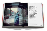 The Assouline book "New York By New York" is open to a spread with a striking image of someone in a long blue coat on a city street, while the opposite page displays a quote about New York City.