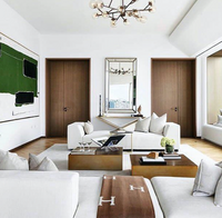 Modern living room with white sofas, brown throw blanket, and glass coffee table featuring the Tizo Lucite Book Stand. Abstract green and black artwork on the wall, two wooden doors, window seat with cushions, and a contemporary chandelier above.