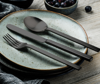 A Mepra Stile Oro Nero 5 Piece Setting—matte black flatware—rests on a light blue ceramic plate with a dark rim on a wooden table. A bowl of blueberries is in the background.