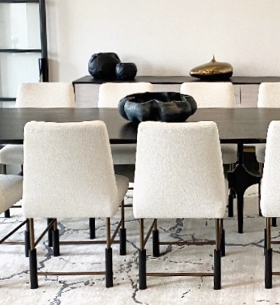 A modern dining room with six white upholstered chairs arranged around a dark rectangular table. Decorative black and gold objects, including the handmade Timna Bowl Blue by L’Objet—evoking treasures from King Solomon’s Mines—are displayed on a sideboard against a white wall. A white patterned rug covers the floor.