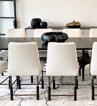 A modern dining room with six white upholstered chairs arranged around a dark rectangular table. Decorative black and gold objects, including the handmade Timna Bowl Blue by L’Objet—evoking treasures from King Solomon’s Mines—are displayed on a sideboard against a white wall. A white patterned rug covers the floor.