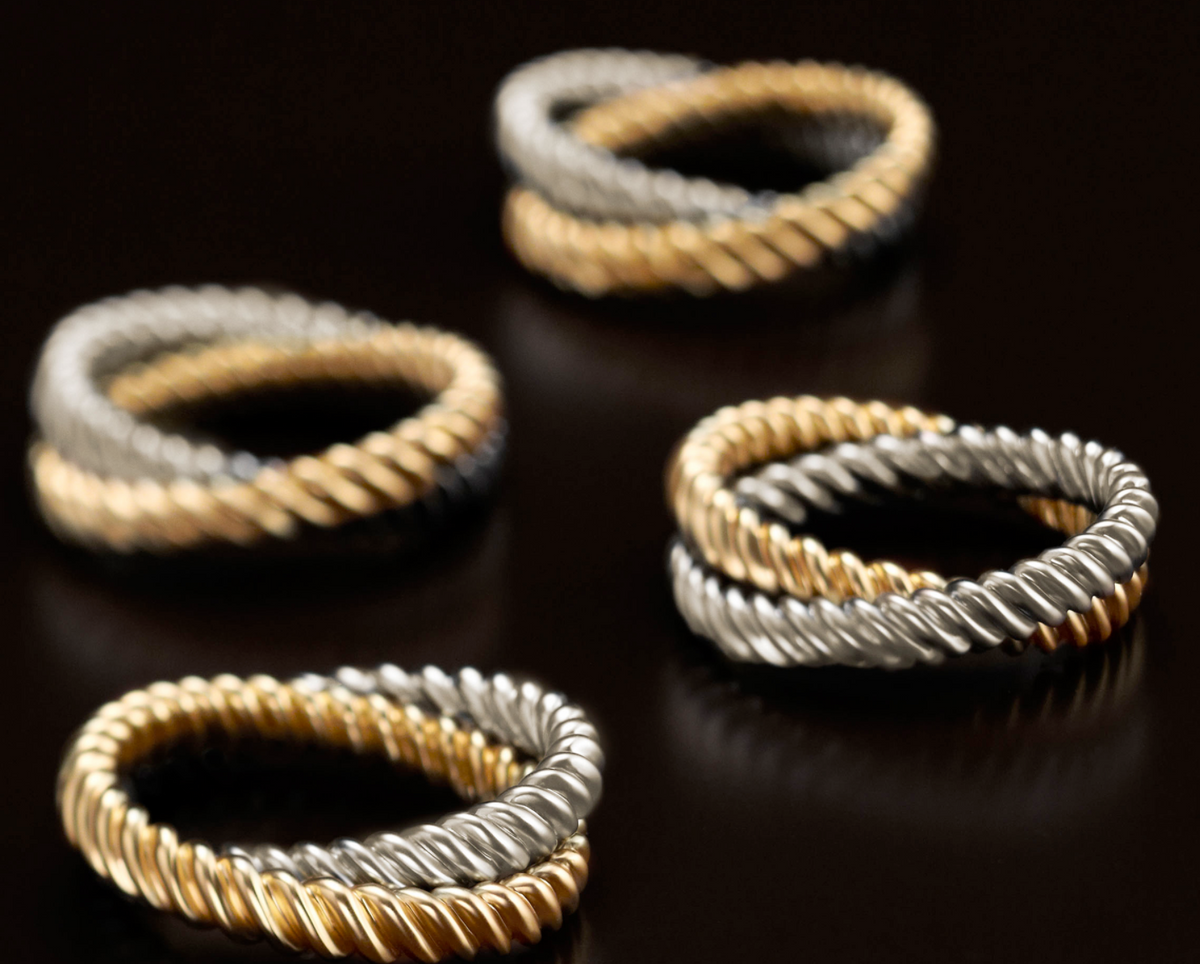 The Deco Twist Napkin Ring Set of 4 by L'Objet, crafted from interwoven gold and silver metals with a 24K gold plating, is displayed on a dark reflective surface—two rings in focus and two softly blurred—a luxurious gift idea.