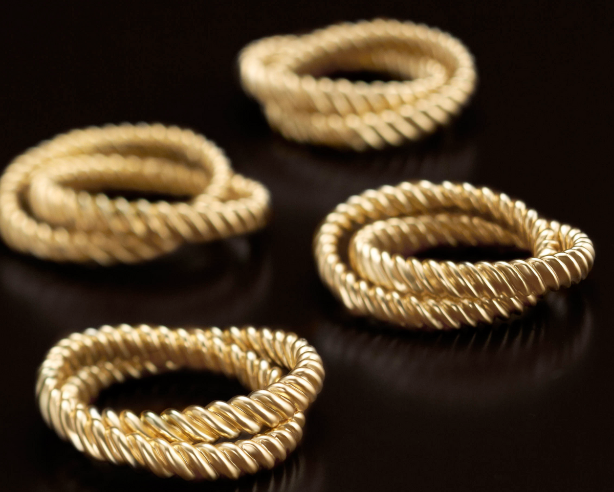 The L'Objet Deco Twist Napkin Ring Set of 4 features twisted 24K gold-plated rings with a rope-like texture, arranged on glossy black for a luxurious, elegant look—ideal as napkin rings or in a luxury gift box.