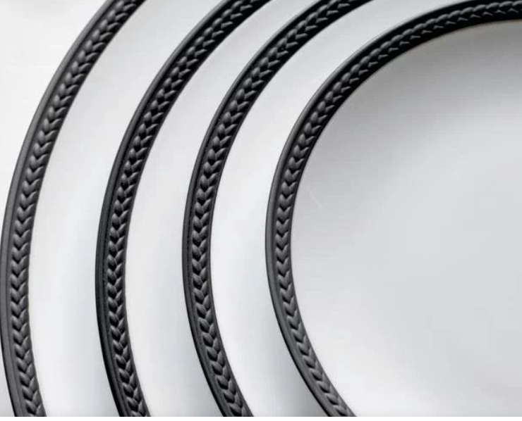 Three Soie Tressée Black porcelain plates by L'Objet, featuring dark, braided-style rims and textured edges, are arranged diagonally on a light background, creating a striking visual motif.