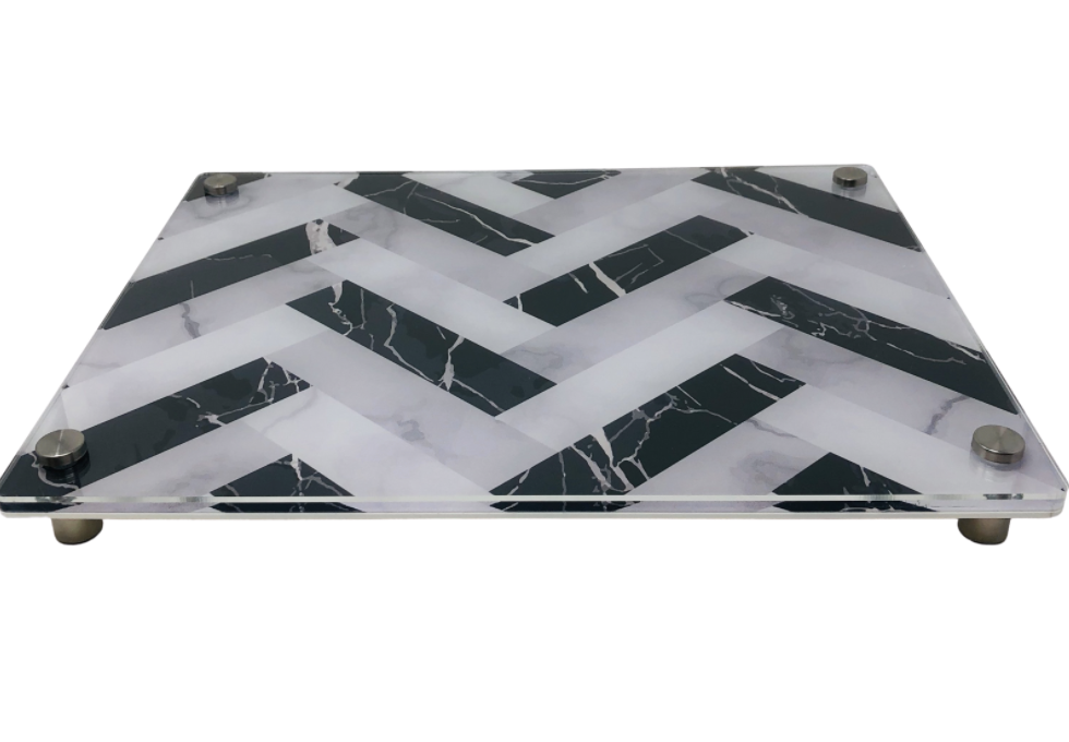 The Eye Candy Raised Serving Board Marble features a rectangular glass surface with a black and white chevron marble pattern, elevated by four metal feet at each corner.