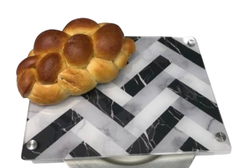 A loaf of braided bread rests on Eye Candy's Raised Serving Board Marble, which features a black and white herringbone marble pattern with rounded metal corners, set against a white background.