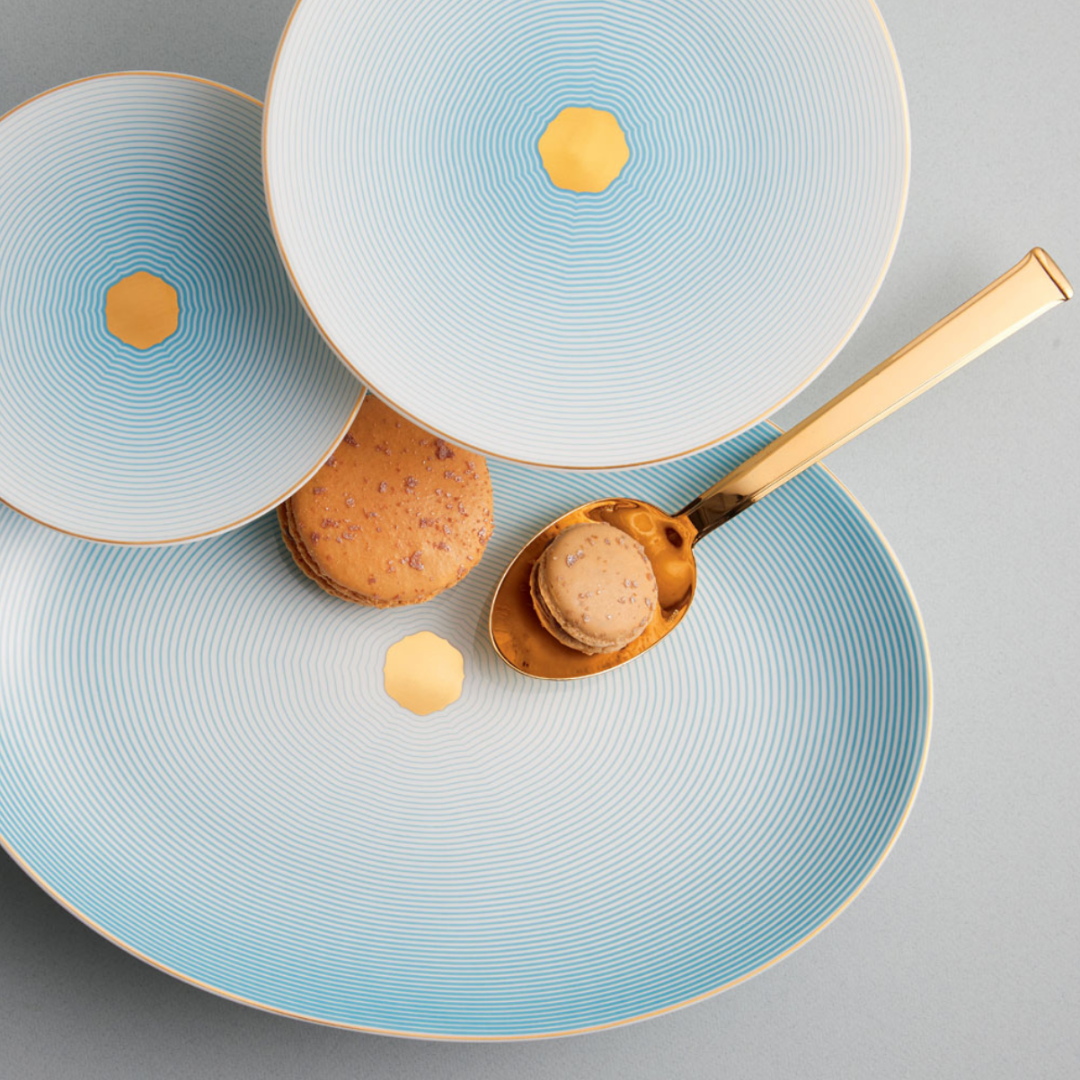 Three Aura Dinnerware plates by Raynaud, featuring blue and white patterns with gold hexagons and scalloped details, overlap elegantly. Two macarons—one on a gold spoon—rest on the plates against a light grey background.
