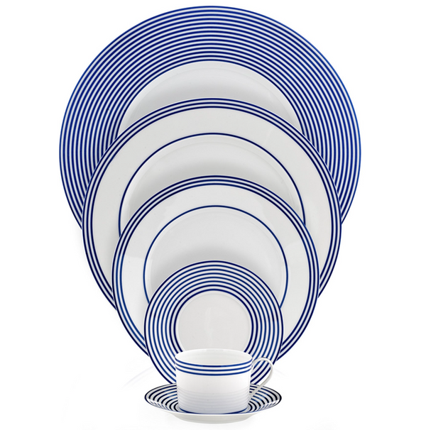The Latitudes Dinnerware by Royal Limoges features handmade white plates and a cup, each adorned with concentric blue rings. The pieces are stacked by size, with the matching cup and saucer displayed at the front.