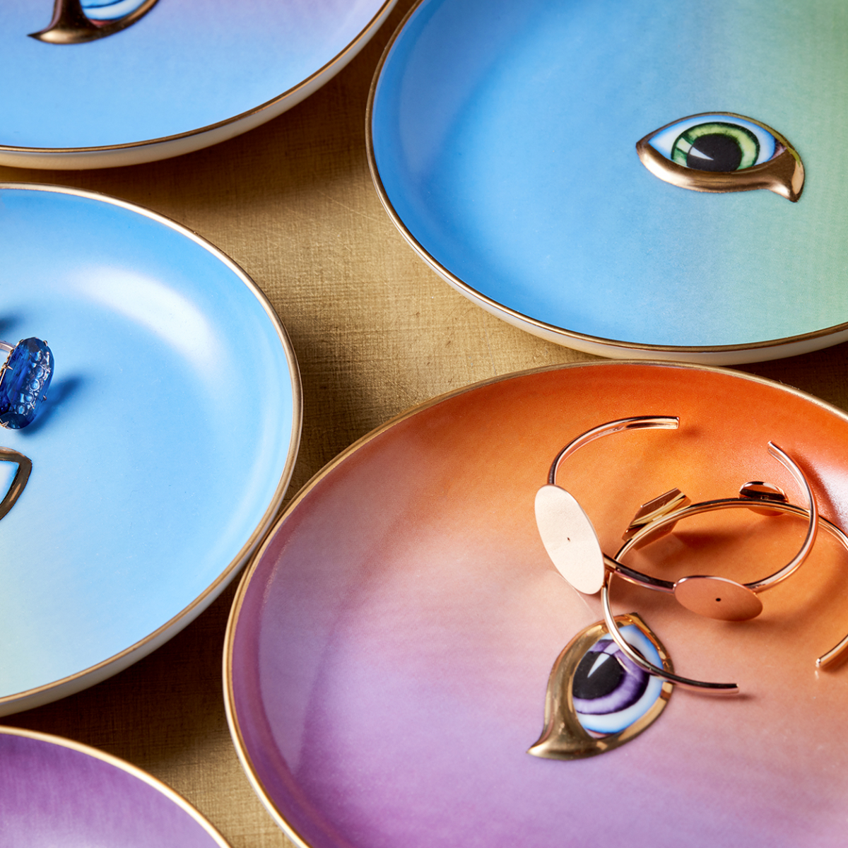 The L’Objet Lito Canape Plate Set of 4 features colorful porcelain plates, each with a painted eye at the center and 24K gold accents, artfully displayed on a beige surface with some holding jewelry for an eclectic touch.