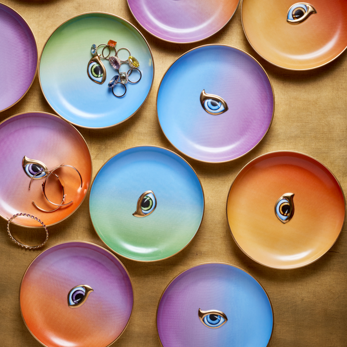 A set of four colorful L’Objet Lito Canape Plates, each with a central eye illustration and 24K gold accents, are displayed on a golden surface; some hold rings and bracelets while others are empty.