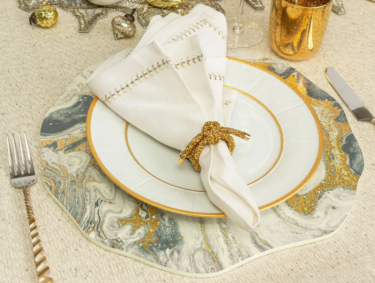Elegant table setting with a Kim Seybert white and gold-rimmed plate, Kim Seybert Herringbone Napkin Gold & Silver in a gold ring, gold flatware, a gold cup, and a marble-patterned placemat on a textured beige tablecloth.
