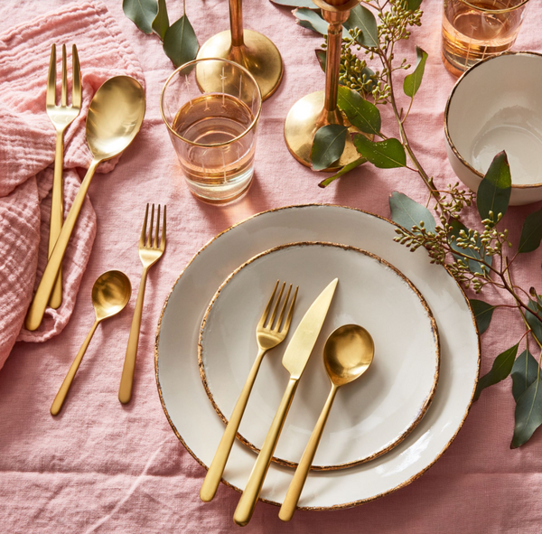 A sophisticated table features Mepra’s Linea 5 Piece Setting – Oro flatware, white plates with gold rims, glassware, brass candlesticks, greenery on a pink tablecloth, and a linen napkin to the left for an elegant touch.