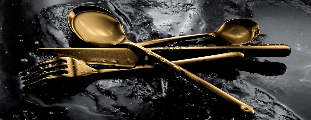 The Mepra Linea 5 Piece Setting - Oro flatware is arranged in a crisscross pattern on a dark, textured surface with water droplets.