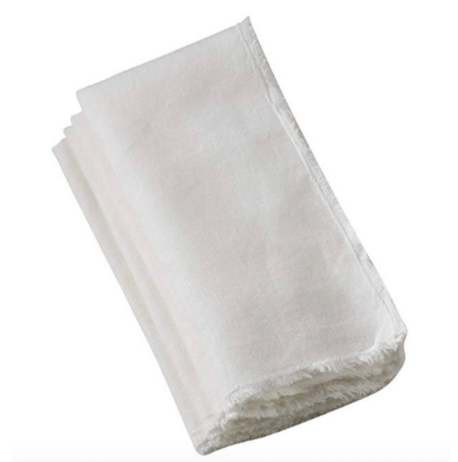 A folded Saro Trading Fringed Stone Washed Napkin in ivory, with subtly frayed edges, is displayed on a plain white background. Set includes 4 napkins.
