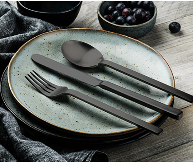 The Mepra Stile Oro Flatware 5 Piece Setting in matte black is arranged on stacked blue ceramic plates, beside a bowl of berries and a gray napkin, all atop a rustic wooden table.