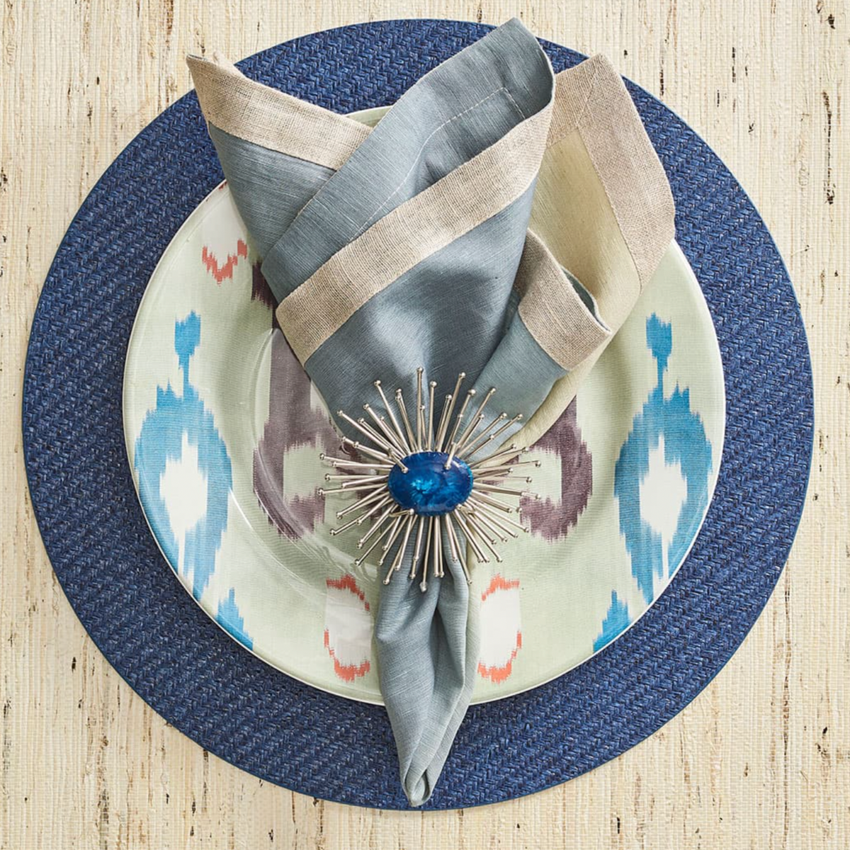 A Kim Seybert Saigon Placemat Navy from the set of 4 holds a patterned blue and gray Ikat plate, topped with a folded gray and beige napkin secured by a starburst napkin ring with a blue center stone.
