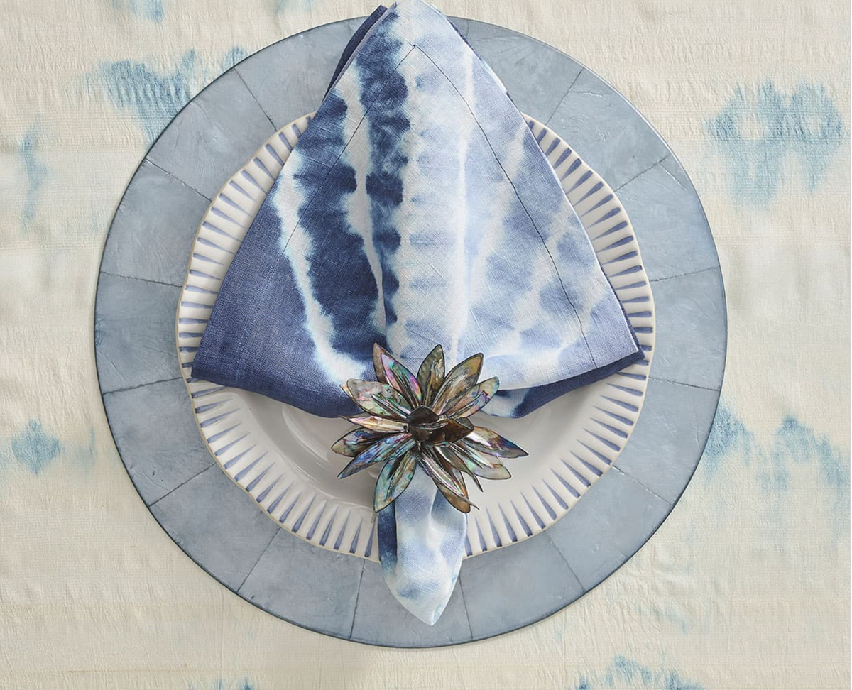 A blue and white tie-dye napkin with a Kim Seybert Shell Flower Napkin Ring, crafted from iridescent river shells, rests on a striped white plate atop a round charger and light blue patterned tablecloth.