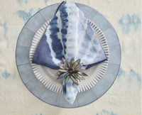 A blue and white tie-dye napkin with a Kim Seybert Shell Flower Napkin Ring, crafted from iridescent river shells, rests on a striped white plate atop a round charger and light blue patterned tablecloth.