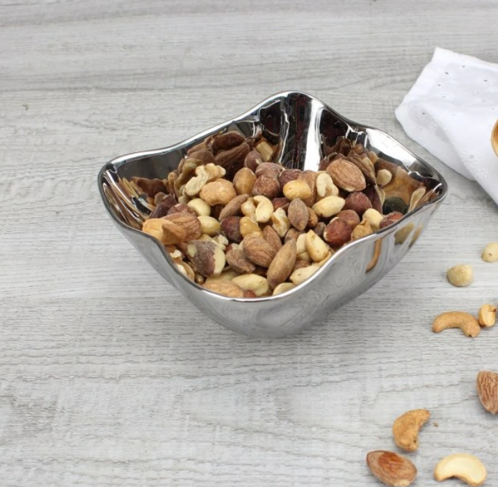 The PAMPA BAY Simple Square Snack Bowl, dishwasher safe for easy cleaning, holds mixed nuts like almonds, cashews, and hazelnuts. Its shiny silver finish sits elegantly on a light wooden surface with scattered nuts and a white cloth in the background.