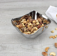 The PAMPA BAY Simple Square Snack Bowl, dishwasher safe for easy cleaning, holds mixed nuts like almonds, cashews, and hazelnuts. Its shiny silver finish sits elegantly on a light wooden surface with scattered nuts and a white cloth in the background.