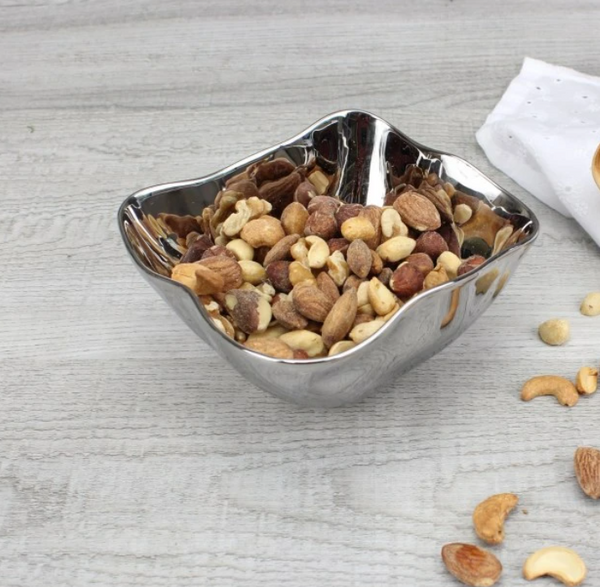 The PAMPA BAY Simple Square Snack Bowl, dishwasher safe for easy cleaning, holds mixed nuts like almonds, cashews, and hazelnuts. Its shiny silver finish sits elegantly on a light wooden surface with scattered nuts and a white cloth in the background.